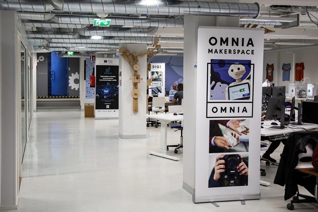 Omnian Makerspace tilaa kuvattuna, edessä tyhjä käytävä ja tolpan edessä Omnian Makerspace -juliste.